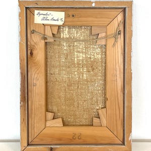May include: The back of a framed painting with a label that reads "Bymaler - Allen Bamble 10." The frame is made of wood and has a light brown finish. The canvas is a natural linen color.