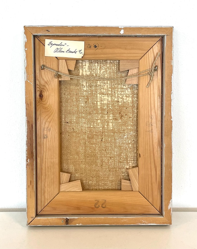 May include: The back of a framed painting with a label that reads "Bymaler - Allen Bamble 10." The frame is made of wood and has a light brown finish. The canvas is a natural linen color.