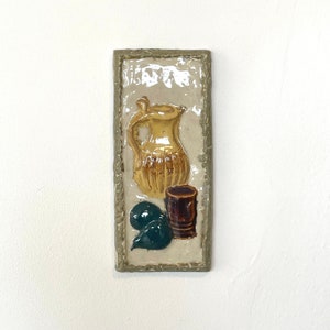 Puede incluir: Un colgante de pared rectangular de cerámica con un diseño en relieve de una jarra amarilla y marrón, una taza marrón y una forma verde oscuro. El fondo es blanquecino con un borde texturizado.