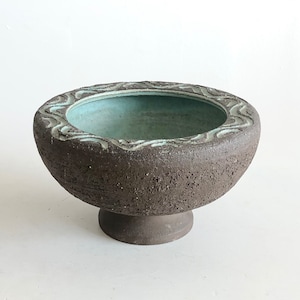 Può includere: Una ciotola in ceramica marrone con una glassa blu chiaro all'interno. La ciotola ha un design ondulato attorno al bordo e si trova su un piedistallo.