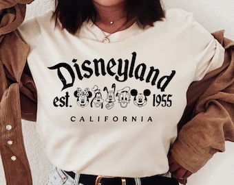 May include: A white t-shirt with a black graphic print that says "Disneyland est. 1955 California" with a line of Mickey Mouse characters. The shirt is being worn by a person with a brown corduroy jacket and white sneakers.