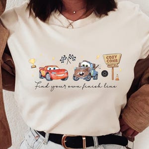 May include: A cream-colored t-shirt featuring a graphic of two cartoon cars, a trophy, checkered flags, and a sign reading Cozy Cone Motel. The text below the graphic reads Find your own finish line. The shirt is worn with a brown corduroy jacket.