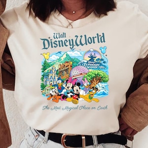 Op de afbeelding: Een cr&egrave;mekleurig t-shirt met een kleurrijke afbeelding van Walt Disney World met personages zoals Mickey Mouse, Minnie Mouse, Donald Duck en Pluto, en de tekst The Most Magical Place on Earth onder de illustratie gedrukt.