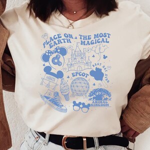 May include: A cream-coloured short-sleeve t-shirt featuring a blue graphic print with the text "Place on Earth the Most Magical" and "Epcot". The design includes illustrations of a castle, a globe, mouse ears, and other park-themed icons.