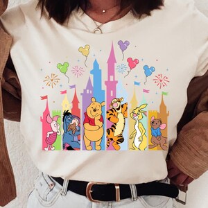 May include: A cream-colored short-sleeve t-shirt featuring a colorful graphic print of a castle silhouette with fireworks and balloons, accompanied by illustrations of Piglet, Eeyore, Winnie the Pooh, Tigger, Roo, and Rabbit standing in a row.