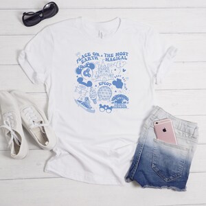 May include: A white short-sleeved t-shirt featuring a blue graphic print with the text "PLACE ON EARTH THE MOST MAGICAL" and illustrations of a castle, a geodesic dome, a tree, a trainer, an ice cream cone, and various park-themed icons.