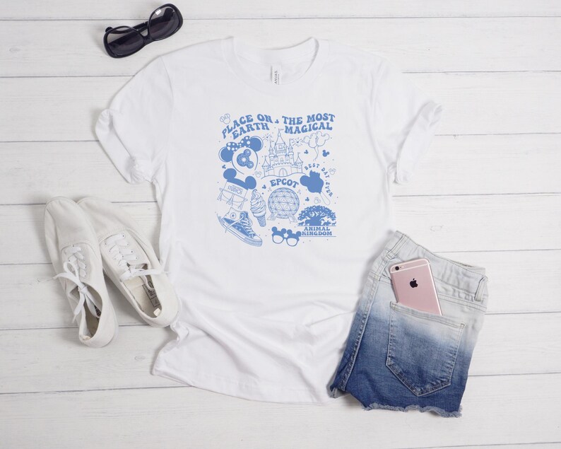 May include: A white short-sleeved t-shirt featuring a blue graphic print with the text "PLACE ON EARTH THE MOST MAGICAL" and illustrations of a castle, a geodesic dome, a tree, a trainer, an ice cream cone, and various park-themed icons.