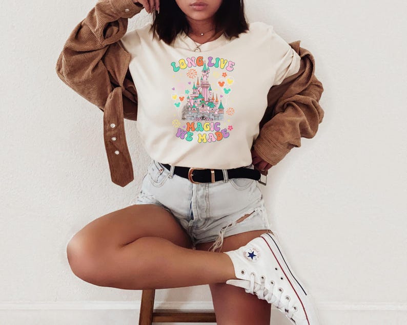 May include: A cream-colored t-shirt featuring a colorful graphic of a castle with the text "LONG LIVE THE MAGIC WE MADE" in a retro font. The design includes small icons like fireworks, stars, and mouse ears in pastel shades of pink, blue, and yellow.
