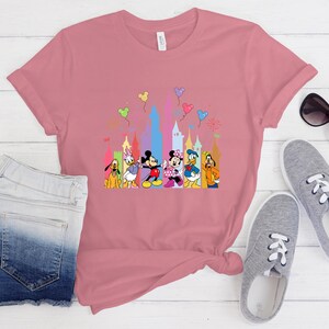 May include: A dusty rose short-sleeved t-shirt featuring a colorful graphic print of Disney characters including Mickey Mouse, Minnie Mouse, Donald Duck, Daisy Duck, Goofy, and Pluto standing in front of a stylized castle with floating balloons.