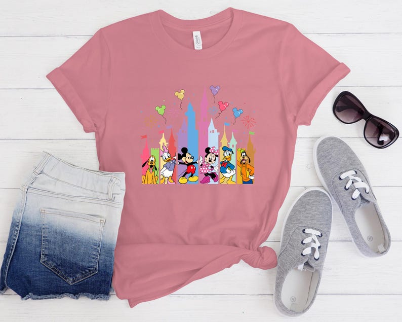 May include: A dusty rose short-sleeved t-shirt featuring a colorful graphic print of Disney characters including Mickey Mouse, Minnie Mouse, Donald Duck, Daisy Duck, Goofy, and Pluto standing in front of a stylized castle with floating balloons.