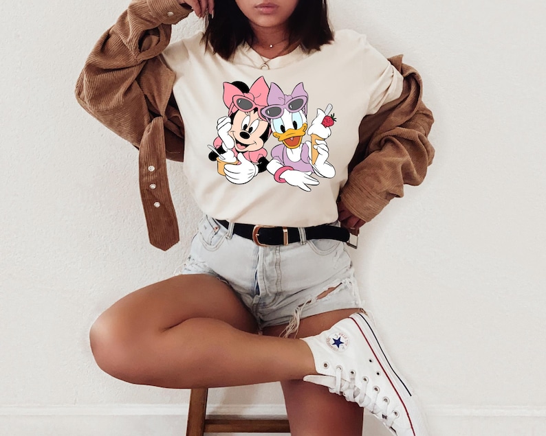 May include: A cream-coloured t-shirt featuring a graphic print of Minnie Mouse and Daisy Duck wearing pink and purple bows and sunglasses, each holding an ice cream cone. The shirt is paired with a brown corduroy button-down shirt and denim shorts.