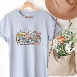 May include: A light blue t-shirt featuring a colourful graphic print of the Magic Kingdom with various park icons and the text Magic Kingdom in a stylised font. The shirt is displayed on a wooden hanger alongside a tan hat and a tan shoulder bag.
