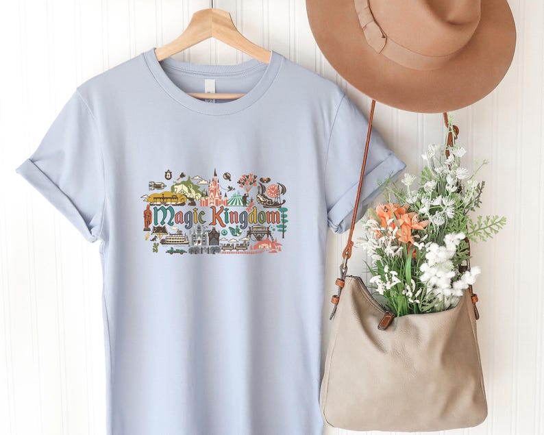 May include: A light blue t-shirt featuring a colourful graphic print of the Magic Kingdom with various park icons and the text Magic Kingdom in a stylised font. The shirt is displayed on a wooden hanger alongside a tan hat and a tan shoulder bag.