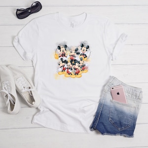 Op de afbeelding: Een wit T-shirt met korte mouwen en een centrale grafische print van meerdere Mickey Mouse-figuren in verschillende houdingen met gele aquarelaccenten. Het shirt wordt getoond met witte sneakers, een zwarte zonnebril en een spijkerbroekshort.