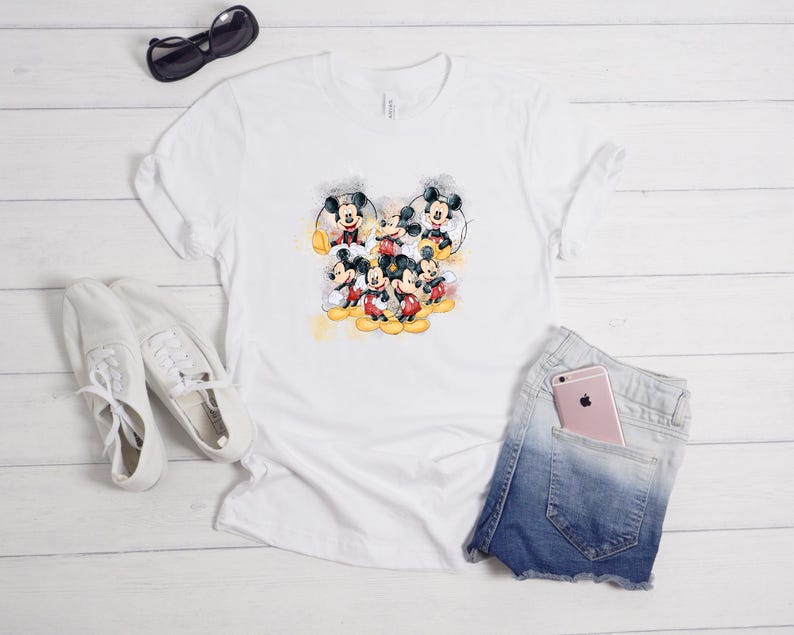 Op de afbeelding: Een wit T-shirt met korte mouwen en een centrale grafische print van meerdere Mickey Mouse-figuren in verschillende houdingen met gele aquarelaccenten. Het shirt wordt getoond met witte sneakers, een zwarte zonnebril en een spijkerbroekshort.