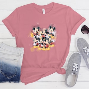 Op de afbeelding: Een oudroze T-shirt met korte mouwen en een centrale grafische print van meerdere Mickey Mouse-figuren in verschillende houdingen, geaccentueerd met gele en oranje spetters en glitters in aquarel-stijl op de voorzijde.