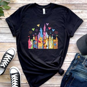 May include: A black short-sleeve t-shirt featuring a colorful graphic print of a castle silhouette with Winnie the Pooh characters including Piglet, Eeyore, Pooh, Tigger, Rabbit, and Roo, accented by small floating Mickey Mouse ear shapes.