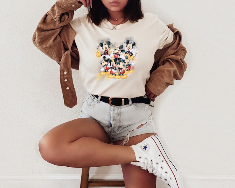 Op de afbeelding: Een cr&egrave;mekleurig T-shirt met korte mouwen en een grafische print van meerdere Mickey Mouse-figuren in verschillende houdingen. Het shirt wordt gecombineerd met een lichtblauwe spijkerbroek, een zwarte riem, een bruin corduroy overhemd en witte sneakers.