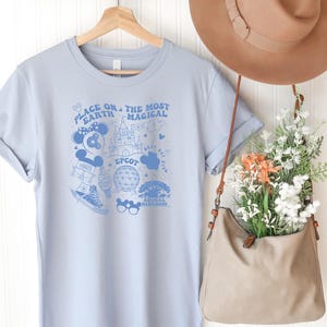 May include: A light blue t-shirt featuring a dark blue graphic print with the text "PLACE ON EARTH THE MOST MAGICAL" and "EPCOT" alongside illustrations of a castle, a geodesic dome, a sneaker, mouse ears, and other park-themed icons.
