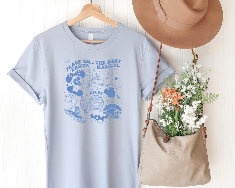 May include: A light blue t-shirt featuring a dark blue graphic print with the text "PLACE ON EARTH THE MOST MAGICAL" and "EPCOT" alongside illustrations of a castle, a geodesic dome, a sneaker, mouse ears, and other park-themed icons.