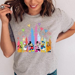 May include: A heather gray short-sleeve t-shirt featuring a colorful graphic print of Disney characters including Mickey Mouse, Minnie Mouse, Donald Duck, Daisy Duck, Goofy, and Pluto standing in front of a stylized castle with floating balloons.