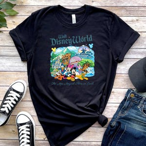 Op de afbeelding: Een zwart t-shirt met korte mouwen en een kleurrijke afbeelding van Walt Disney World-bezienswaardigheden en personages, waaronder Mickey Mouse, met de tekst "Walt Disney World" en "The Most Magical Place on Earth" op de borst.