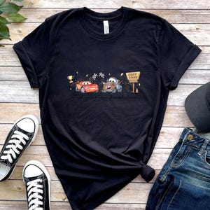 May include: A black short-sleeve t-shirt featuring a graphic print of a red race car and a brown tow truck. The design includes a gold trophy, checkered flags, and a sign that reads Cozy Cone Motel. The shirt has a crew neckline and short sleeves.