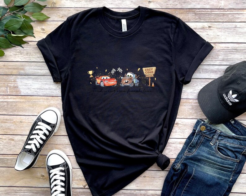 May include: A black short-sleeve t-shirt featuring a graphic print of a red race car and a brown tow truck. The design includes a gold trophy, checkered flags, and a sign that reads Cozy Cone Motel. The shirt has a crew neckline and short sleeves.