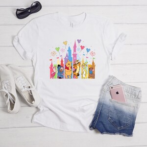 May include: A white short-sleeved t-shirt featuring a colorful graphic of Winnie the Pooh characters standing in front of a castle silhouette with balloons and fireworks. The design includes Piglet, Eeyore, Pooh, Tigger, Rabbit, and Roo.