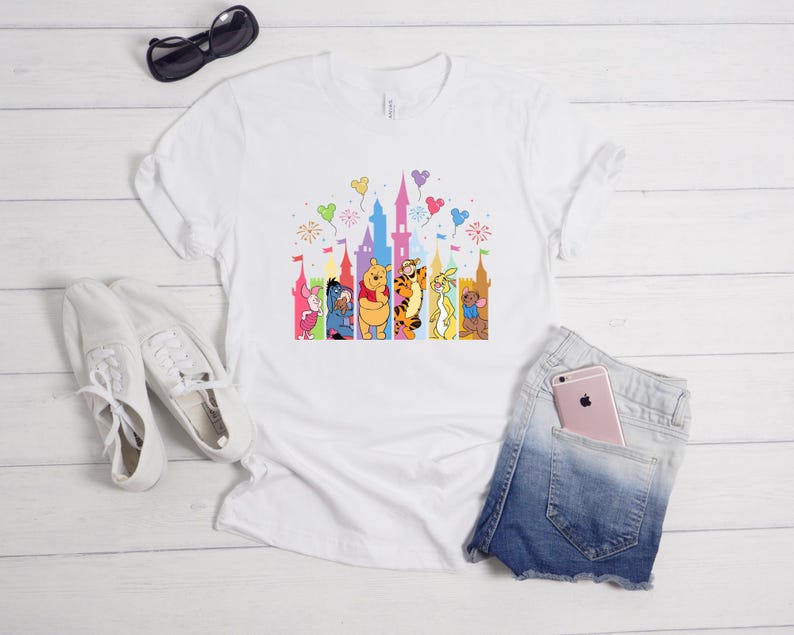 May include: A white short-sleeved t-shirt featuring a colorful graphic of Winnie the Pooh characters standing in front of a castle silhouette with balloons and fireworks. The design includes Piglet, Eeyore, Pooh, Tigger, Rabbit, and Roo.
