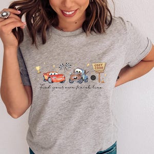 May include: A heather gray t-shirt featuring a graphic print of a red race car and a brown tow truck. The design includes a gold trophy, two checkered flags, a sign reading Cozy Cone Motel, and the text Find your own finish line.