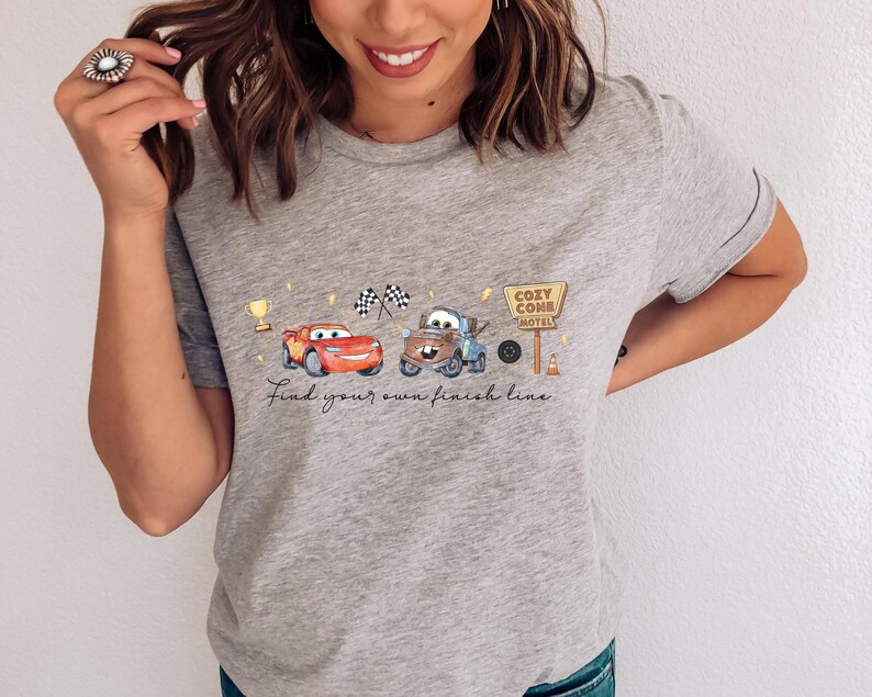 May include: A heather gray t-shirt featuring a graphic print of a red race car and a brown tow truck. The design includes a gold trophy, two checkered flags, a sign reading Cozy Cone Motel, and the text Find your own finish line.