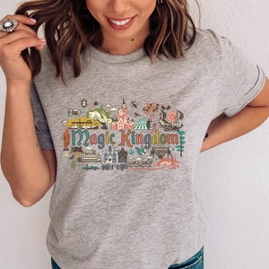 May include: A heather grey short-sleeve t-shirt featuring a colourful graphic print of various theme park attractions and the text Magic Kingdom in a stylised font. The design includes illustrations of a castle, a ship, and other park icons.