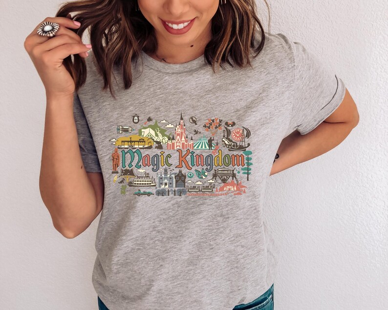 May include: A heather grey short-sleeve t-shirt featuring a colourful graphic print of various theme park attractions and the text Magic Kingdom in a stylised font. The design includes illustrations of a castle, a ship, and other park icons.