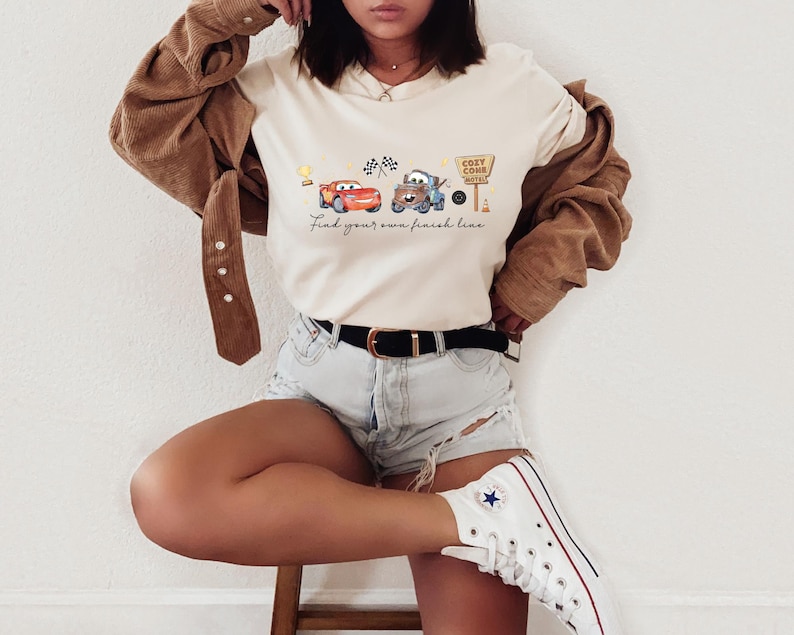 May include: A cream-colored t-shirt featuring a graphic of two cartoon cars, a trophy, checkered flags, and a sign reading Cozy Cone Motel. The text below the graphic reads Find your own finish line. The shirt is worn with a brown corduroy jacket.