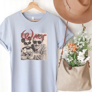 Puede incluir: Una camiseta azul claro con un estampado gr&aacute;fico de Walt Disney y Mickey Mouse con gafas de sol, con la palabra "Walt" en letra roja encima. La camiseta se muestra en una percha de madera junto a un sombrero y un bolso beige con flores.