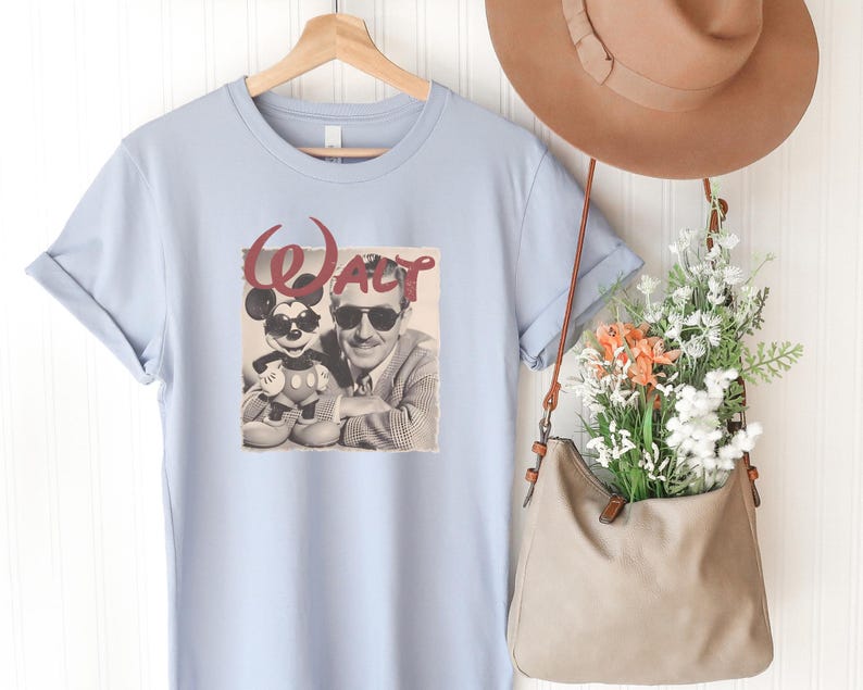 Puede incluir: Una camiseta azul claro con un estampado gr&aacute;fico de Walt Disney y Mickey Mouse con gafas de sol, con la palabra "Walt" en letra roja encima. La camiseta se muestra en una percha de madera junto a un sombrero y un bolso beige con flores.