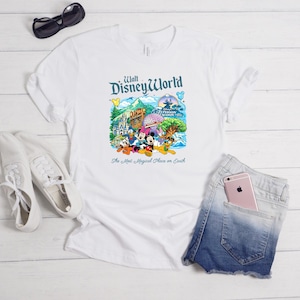 Op de afbeelding: Een wit t-shirt met korte mouwen en een kleurrijke afbeelding van Walt Disney World met personages zoals Mickey Mouse, Minnie Mouse, Donald Duck en Pluto, inclusief de tekst "Walt Disney World" en "The Most Magical Place on Earth."
