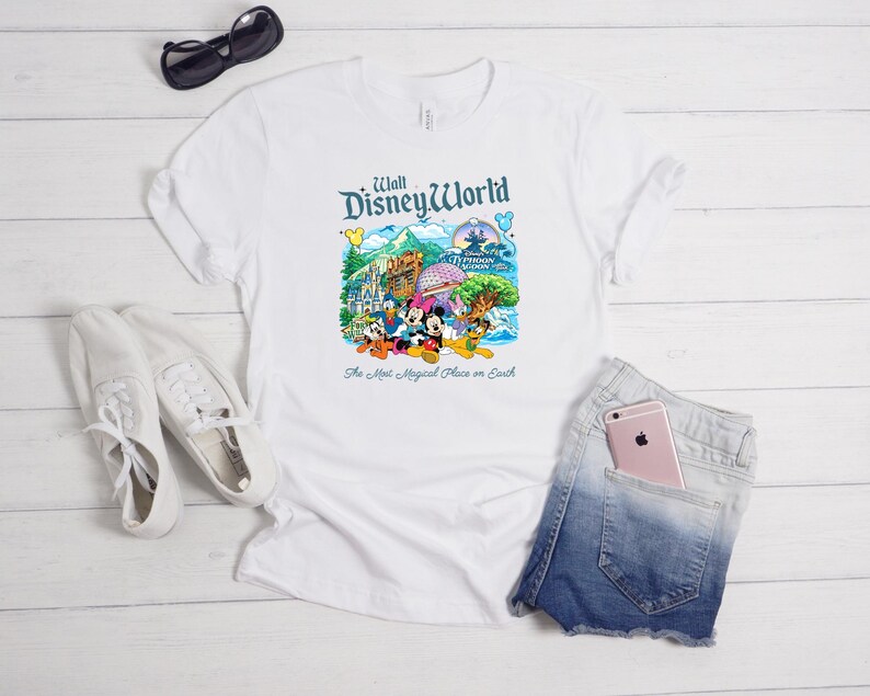 Op de afbeelding: Een wit t-shirt met korte mouwen en een kleurrijke afbeelding van Walt Disney World met personages zoals Mickey Mouse, Minnie Mouse, Donald Duck en Pluto, inclusief de tekst "Walt Disney World" en "The Most Magical Place on Earth."