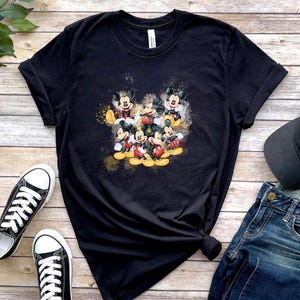 Op de afbeelding: Een zwart t-shirt met korte mouwen en een centrale grafische print van meerdere Mickey Mouse-figuren in verschillende houdingen, uitgevoerd in een aquarel-stijl met gele en oranje accenten op de donkere stoffen achtergrond.