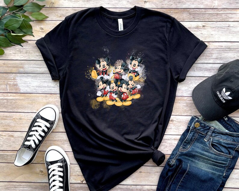 Op de afbeelding: Een zwart t-shirt met korte mouwen en een centrale grafische print van meerdere Mickey Mouse-figuren in verschillende houdingen, uitgevoerd in een aquarel-stijl met gele en oranje accenten op de donkere stoffen achtergrond.