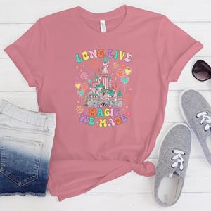 May include: A dusty rose short-sleeve t-shirt featuring a graphic of a castle with the text "LONG LIVE" and "ALL THE MAGIC WE MADE" in colorful, retro-style lettering, surrounded by illustrations of fireworks and balloons.