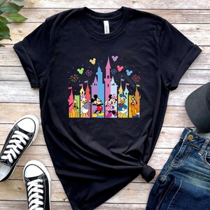May include: A black short-sleeved t-shirt featuring a colorful graphic print of a castle silhouette with fireworks and Disney characters including Pluto, Daisy Duck, Mickey Mouse, Minnie Mouse, Donald Duck, and Goofy standing in front.