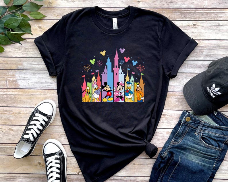 May include: A black short-sleeved t-shirt featuring a colorful graphic print of a castle silhouette with fireworks and Disney characters including Pluto, Daisy Duck, Mickey Mouse, Minnie Mouse, Donald Duck, and Goofy standing in front.