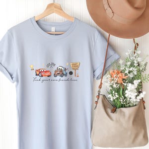 May include: A light blue t-shirt featuring a graphic of two cartoon cars, a checkered flag, and a sign that reads Cozy Cone Motel. Below the cars, the text reads Find your own finish line. The shirt is displayed on a wooden hanger.