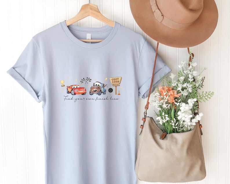 May include: A light blue t-shirt featuring a graphic of two cartoon cars, a checkered flag, and a sign that reads Cozy Cone Motel. Below the cars, the text reads Find your own finish line. The shirt is displayed on a wooden hanger.