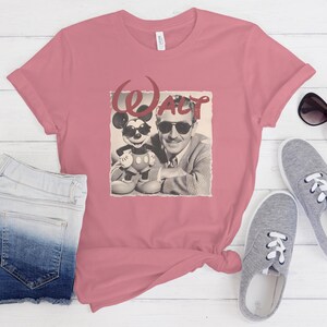 Puede incluir: Una camiseta de manga corta color rosa empolvado con un estampado gr&aacute;fico de Walt Disney y Mickey Mouse, ambos con gafas de sol. El texto "WALT" est&aacute; impreso en una fuente roja estilizada sobre las figuras.