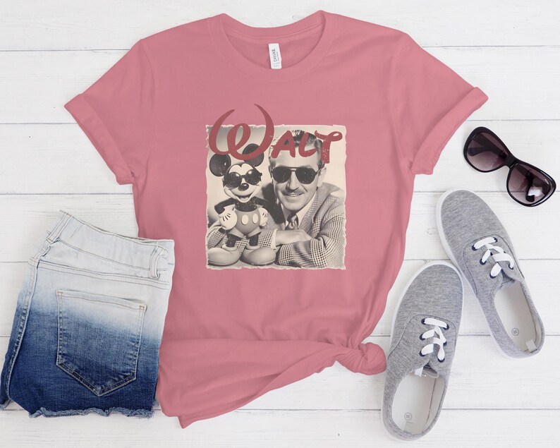 Puede incluir: Una camiseta de manga corta color rosa empolvado con un estampado gr&aacute;fico de Walt Disney y Mickey Mouse, ambos con gafas de sol. El texto "WALT" est&aacute; impreso en una fuente roja estilizada sobre las figuras.