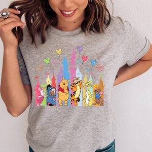 May include: A heather gray short-sleeve t-shirt featuring a colorful graphic print of Winnie the Pooh characters including Piglet, Eeyore, Pooh, Tigger, Rabbit, and Roo standing in front of a stylized castle silhouette with floating balloons.
