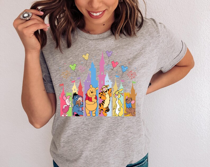 May include: A heather gray short-sleeve t-shirt featuring a colorful graphic print of Winnie the Pooh characters including Piglet, Eeyore, Pooh, Tigger, Rabbit, and Roo standing in front of a stylized castle silhouette with floating balloons.